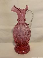 Front view showing textured cranberry glass body, clear applied handle, ruffled spout, and pedestal base.
