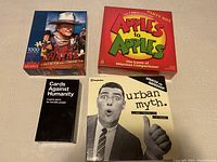 Four items displayed on a surface: John Wayne 1000 piece puzzle, Apples to Apples party box, Cards Against Humanity black box, and Urban Myth game box.