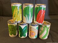 Seven hermetically sealed metal seed canisters with colorful vintage labels showing various vegetables. They are arranged standing and stacked.