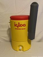 Front view of yellow Igloo drinking water cooler with red lid and attached gray cup dispenser