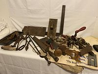 Wide view of vintage tools including rustic metal pieces, hand saw, and wooden handled tools laid out on a white fabric
