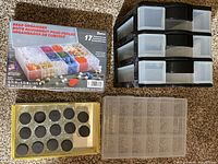 Photo shows 3 plastic organizers stacked on the right, and a box of 14 glass bottles with black lids to the left on carpeted floor.