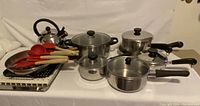 Full assortment photo showing two silicone spatulas, wooden spoons, a whistling tea kettle, several stainless steel pots and pans, drying rack and grill topper.