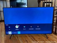 Frontal view of powered-on TV showing home screen with sources menu and V-shaped feet on wood floor