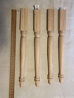 Four unfinished wooden table legs laid out side by side showing the turned and carved design and square tops.
