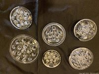 Top-down view of six round clear glass flower frogs arranged on dark surface, varying sizes and multiple holes each.