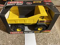 Front-facing photo showing the yellow steel toy dump truck inside original black Tonka Mighty Dump box with clear window. The truck is visible through the window, and the box shows some wear including scuffs and stickers.
