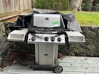 Front view of stainless steel Napoleon propane BBQ grill showing four control knobs and two side shelves with a cover draped over the back.