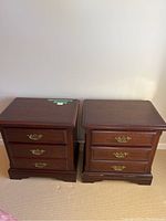 Photo showing two matching wooden bedside tables side by side against a wall, each with three drawers and ornate gold-colored handles.
