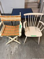 Director chair with floral fabric and white wooden frame, occasional chair in white wood, and blue card table in garage setting.