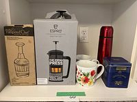 Photo shows the boxed Espro P5 coffee press, boxed Pampered Chef chopper, four mugs including a floral pattern one, small bowl, red metallic thermos, and blue Golden Valley tin container on a shelf.