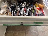 Top view of drawer filled with assorted cutlery and kitchen utensils including spoons, knives, peelers, and other tools.