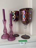 Photo showing two purple glass candle holders with purple taper candles next to two large mosaic-style wine glass candle holders on a white shelf.
