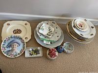 Photo showing assortment of decorative and commemorative plates, cake stand, small thimbles, and figurine items arranged in groups on a carpeted floor.