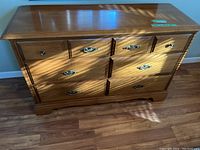 Front view of wooden dresser showing six drawers with decorative metal handles and polished finish. Visible signs of surface wear and used condition.