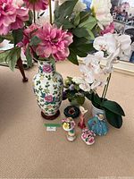 Photo showing tall ceramic vase with floral pattern, artificial pink and white flowers, small decorative porcelain pieces including floral arrangements and figurine