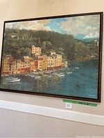 Framed canvas painting showing a coastal village, colorful buildings, and boats in a bay under a partly cloudy sky.