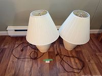 Two matching beige ceramic table lamps with tall white pleated fabric shades on wooden floor against gray wall.