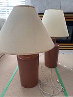 Two table lamps with cylindrical reddish-brown ceramic bases and cone-shaped cream fabric lampshades, cords visible on carpeted floor.