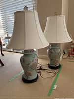 Photo of two matching Asian inspired ceramic lamps with wood bases on carpeted floor near a window, showing shades and cords.