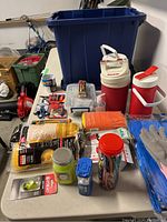 Photo of table top with many outdoor items arranged including coolers, fishing gear, rope, gloves, flasks, small parts containers, and a blue tarp.
