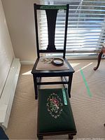 Full view of small dark wooden rocking chair with floral needlepoint seat, matching footstool with green floral needlepoint upholstery in front, sunlight coming from window.