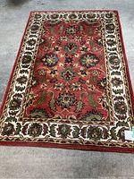 Full view of the red handmade rug with floral patterns and cream border on concrete floor.