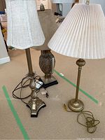 Three ornate table lamps together on a beige carpet, showing details of their bases and fabric shades. One lamp has a fluted gold-toned metal base with a white pleated shade; another lamp has a linished brass tall base and beige bell-shaped shade; third lamp has a darker bronze urn-shaped base.