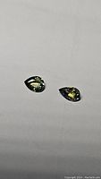 Two pear-shaped yellow green blue tone sapphire gemstones lying flat on a white surface showing their shape and color.