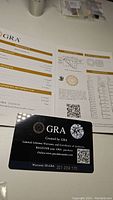 Photos of the GRA certificate, grading report showing the moissanite details including 1 carat weight, D color, VVS1 clarity, and cushion cut shape. Included GRA warranty card with unique ID.