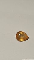 Close-up photo of a single pear-shaped faceted deep golden yellow sapphire gemstone on a plain light background.