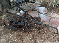 Full view of antique iron and wood hand plow displaying the long wooden handles and iron frame resting on soil with flagstone background.