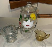 Full view of three glass kitchen items on counter: Bartlett Collins floral cookie jar, Pyrex measuring cup, amber depression glass creamer