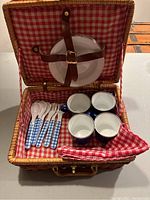 Open wicker picnic basket showing arrangement of four blue enamelware plates secured with leather straps on lid, four blue enamelware cups, and plastic utensils with blue gingham check handles inside basket lined with red gingham check fabric.