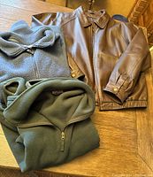 Three men's jackets displayed on a wood table: brown leather bomber, green fleece half-zip, green fleece full zip.