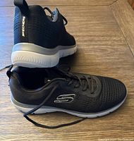 Side and top angle showing black Skechers sneaker with white sole, breathable mesh upper and Skechers branding on side and heel tab.