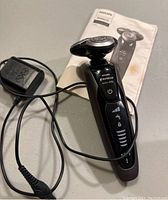 Front view of black Philips Norelco Series 9006 shaver with charger cable and instruction manual beneath.