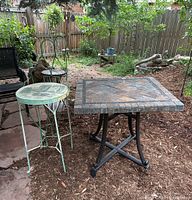 Outdoor setting showing the square wrought iron patio table with multi-colored stone tile top and adjacent painted iron chair with circular seat.