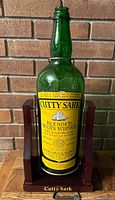 Full view of the vintage one gallon green glass Cutty Sark whisky bottle in the wooden swing stand against a brick wall.