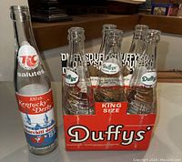 Six vintage soda bottles displayed including five Duffys pop bottles in original close-up red Duffys cardboard carrier and one separate 1974 RC Cola Churchill Downs commemorative bottle.
