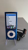 Front view of the blue Apple iPod Nano screen showing the main menu with options including Video Camera, Music, and Podcasts. The charging/data cable is visible behind the device.