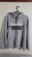 Grey hoodie with New York City graphic print on front hanging on black hanger against white wall.