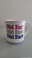 Front view of white ceramic mug with 'Old Fart' repeated three times in red, gray, and blue lettering.