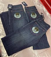 Three pairs of folded dark wash jeans with visible skull embroidery on back pockets, new with tags attached.