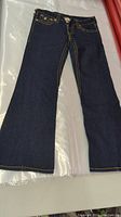 Full view of the front side of the women's True Religion bootcut denim jeans laid flat showing the dark blue color and bootcut pant legs with yellow stitching.