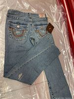 Full view of light blue denim jeans laid flat showing back pockets with distinctive stitching and tags attached.