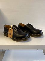 Side angled view of the pair of black leather Dr. Martens shoes with original tag attached, showing smooth leather and yellow stitching.