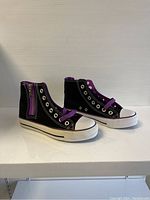 Pair of black and purple high-top sneakers with white rubber soles and toe caps, shown from a side angle to highlight front and side zippers and laces.