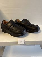 Side angle view of a pair of men's black leather shoes with Velcro straps, placed on white surface.