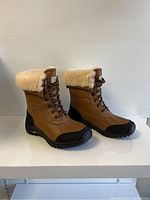 Pair of brown UGG Adirondack II ladies winter boots with black soles and fluffy sheepskin collars photographed from left side angle.